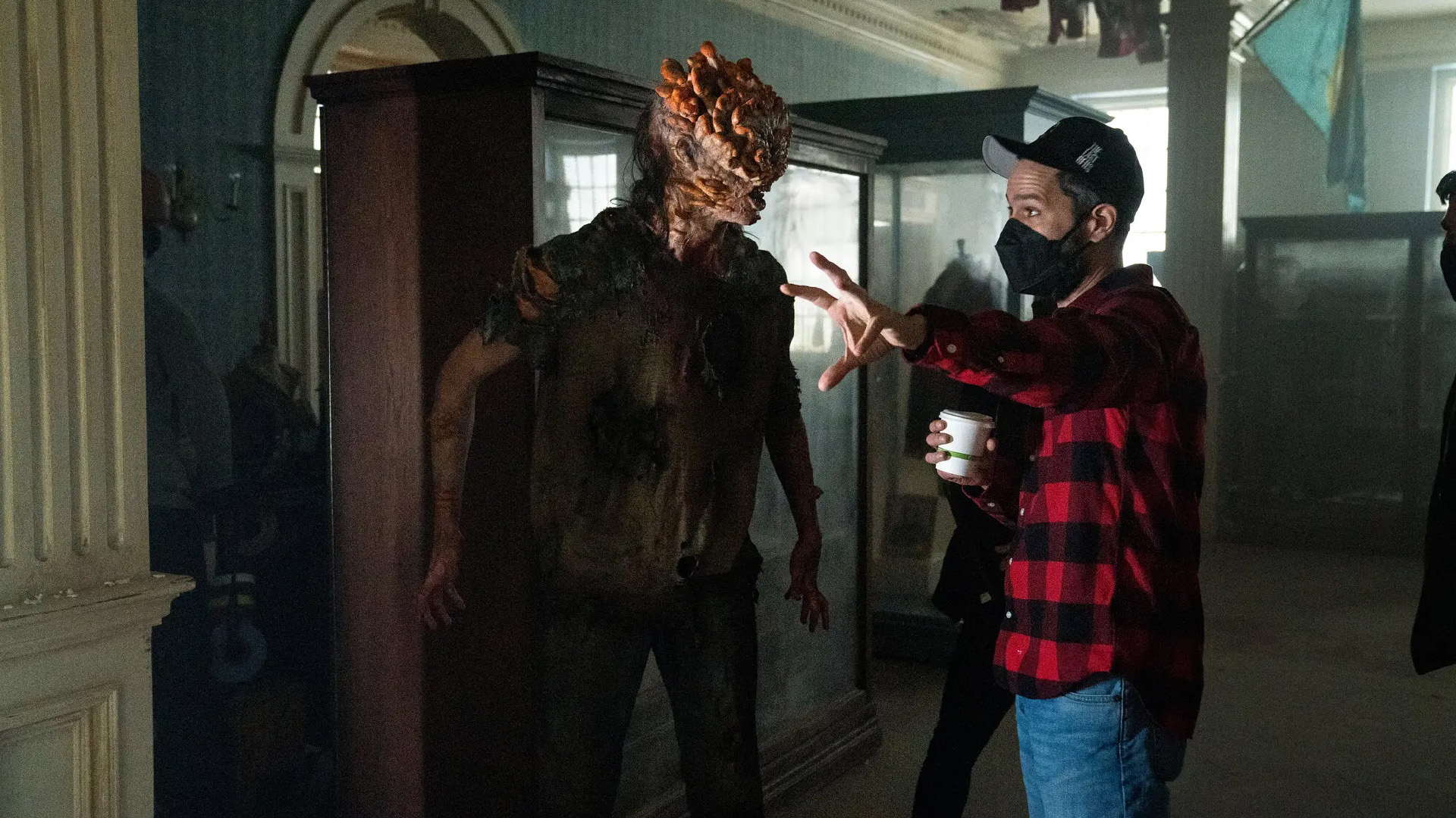 Photo des coulisses de la série The Last of Us (S01E02). Neil Druckmann, vêtu d'une casquette, d'une chemise rouge à carreaux noirs et d'un masque pour se protéger du covid, donne des instructions à un·e comédien·ne grimmé en claqueur sur le tournage.