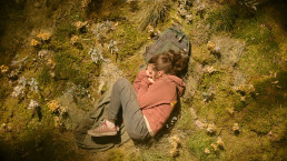 Vue en plongée sur Ellie, jouée par Bella Ramsey. Elle est recroquevillée en position fœtale pour dormir, installée sur un lit d'herbe et de mousse, la tête posée sur son sac à dos tandis que la lumière du jour l'éclaire.