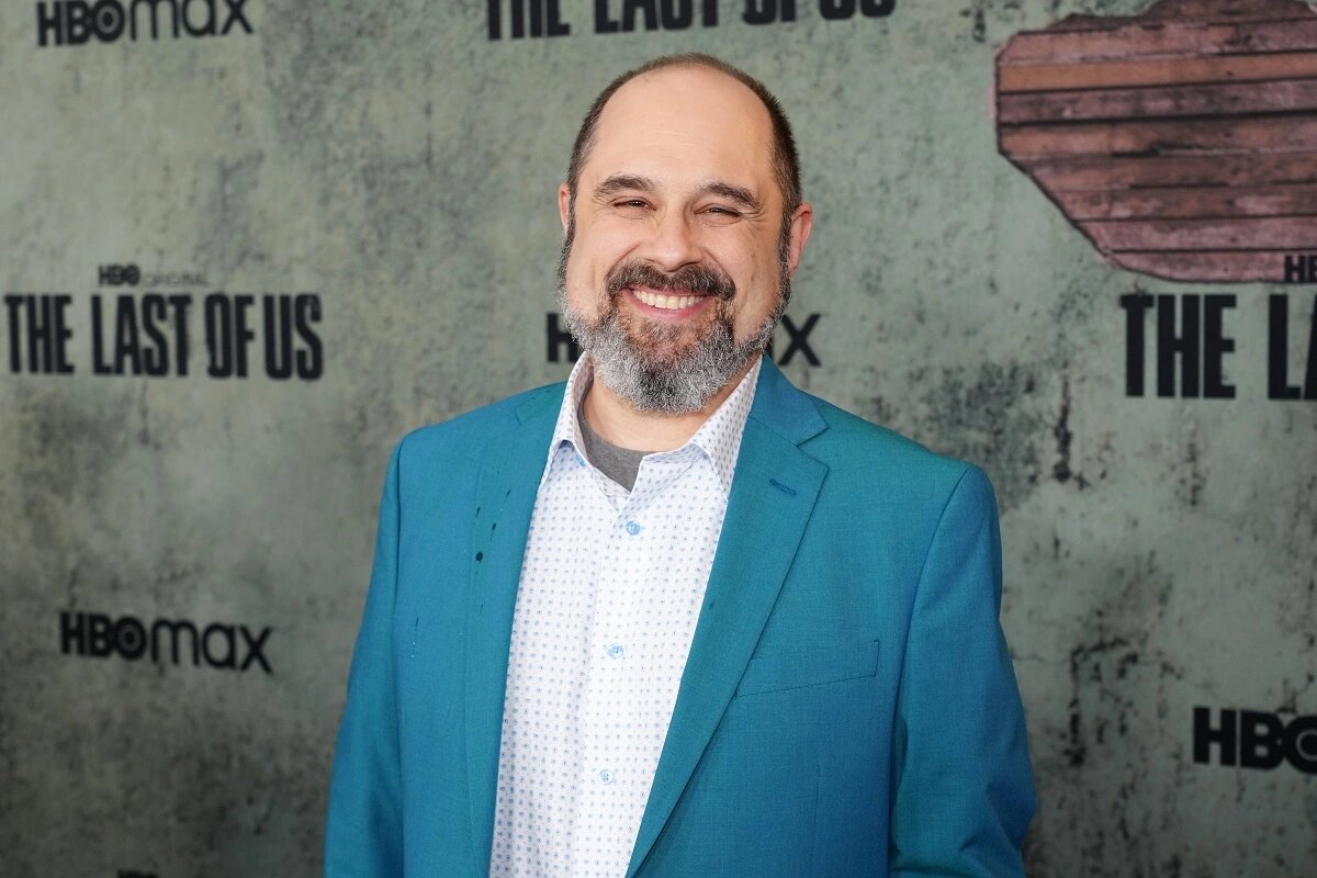 Craig Mazin with a big smile and a costume mischievous in front of a dilapidated wall on which The Last of Us (HBO) series logo has been plastered several times.