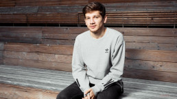 Portrait de Kantemir Balagov, assis sur des gradins en bois. Il porte un pantalon noir et un sweat Adidas.