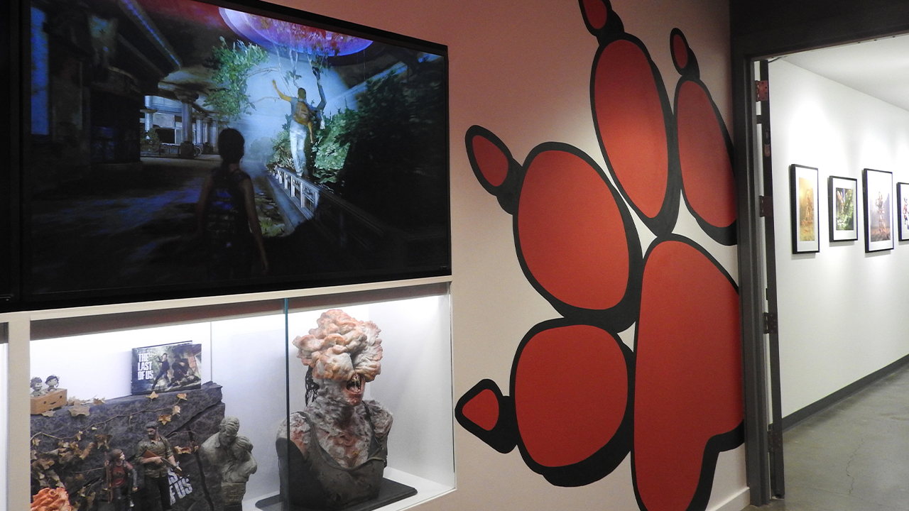 Photo d'un couloir des bureaux de Naughty Dog. Un autocollant géant du logo du studio - une empreinte rouge de patte de chien - est collé au mur, à côté dun écran sur lequel est diffusé le jeu vidéo The Last of Us, et d'une vitrine dans laquelle sont exposés des statuettes d'infectés.