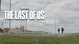 Plan large, en extérieur, d'une route bordée par de l'herbe. Au loin, les silouhettes de Ellie et Joel poursuivent leur route, cote à cote. (The Last of Us HBO, saison 1, épisode 5)